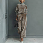 A woman wears a taupe velour high-neck long-sleeve top and matching pleated wide-leg pants against a gray wall.