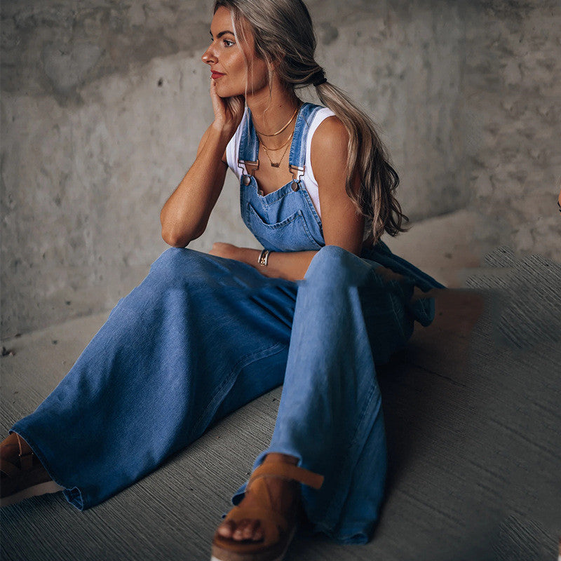 Woman wearing a white tank top under wide-leg blue denim overalls, paired with brown strappy sandals.