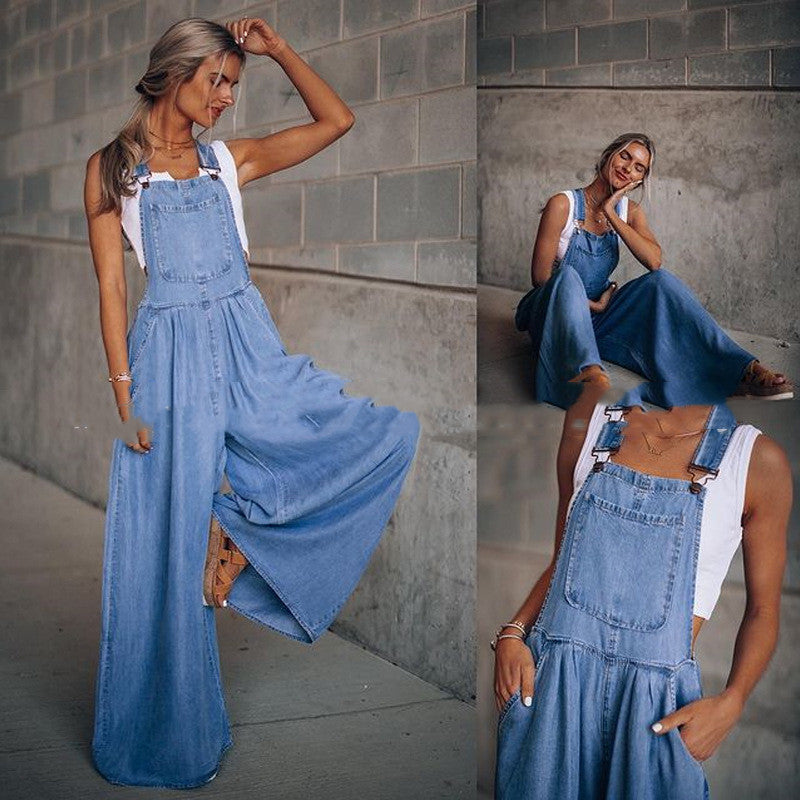 A woman wears a light-wash denim overall jumpsuit with a wide-leg silhouette over a white tank top.