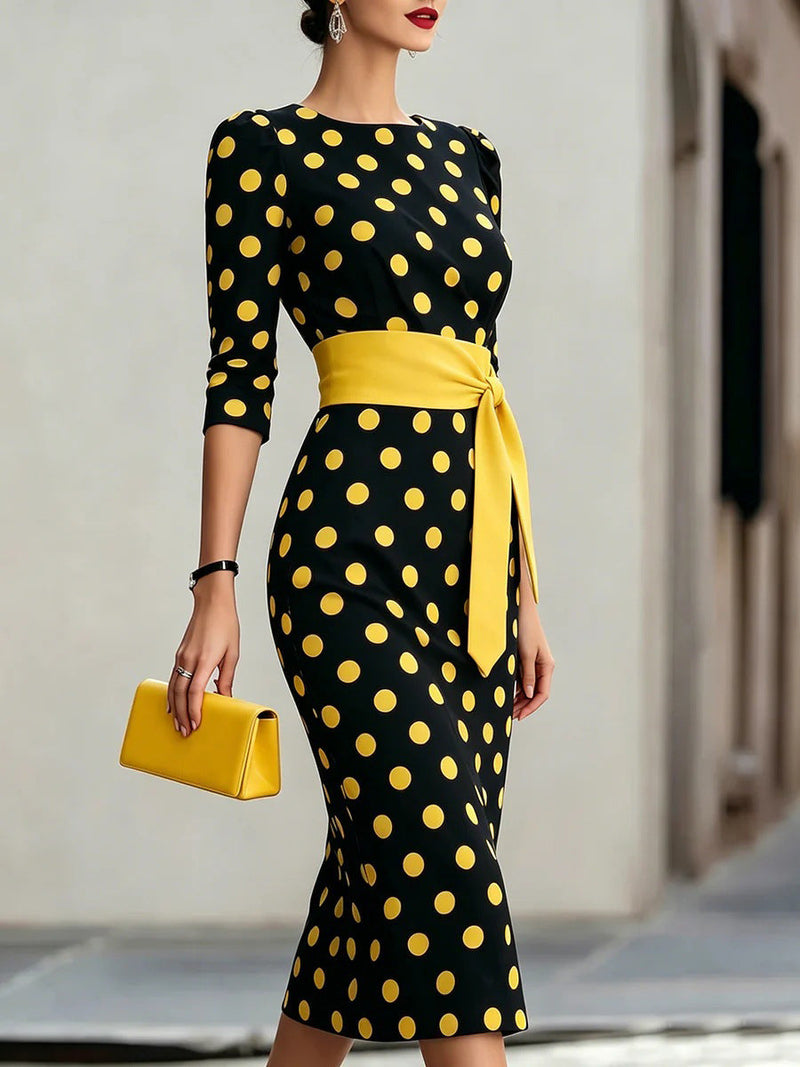 Woman in a black midi dress with yellow polka dots, featuring three-quarter sleeves, a yellow sash, and a matching bag.