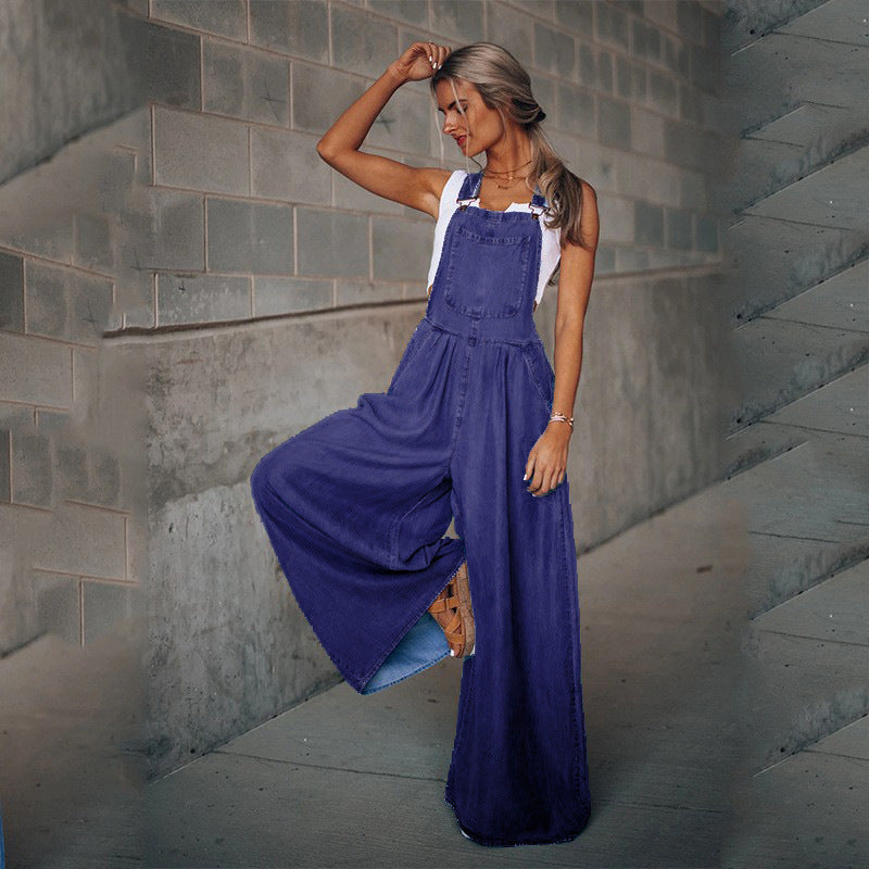 A woman wears loose-fitting deep blue denim overalls with wide legs over a simple white tank top.