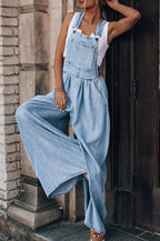 A person wearing light-wash denim overalls with wide legs over a white tank top.