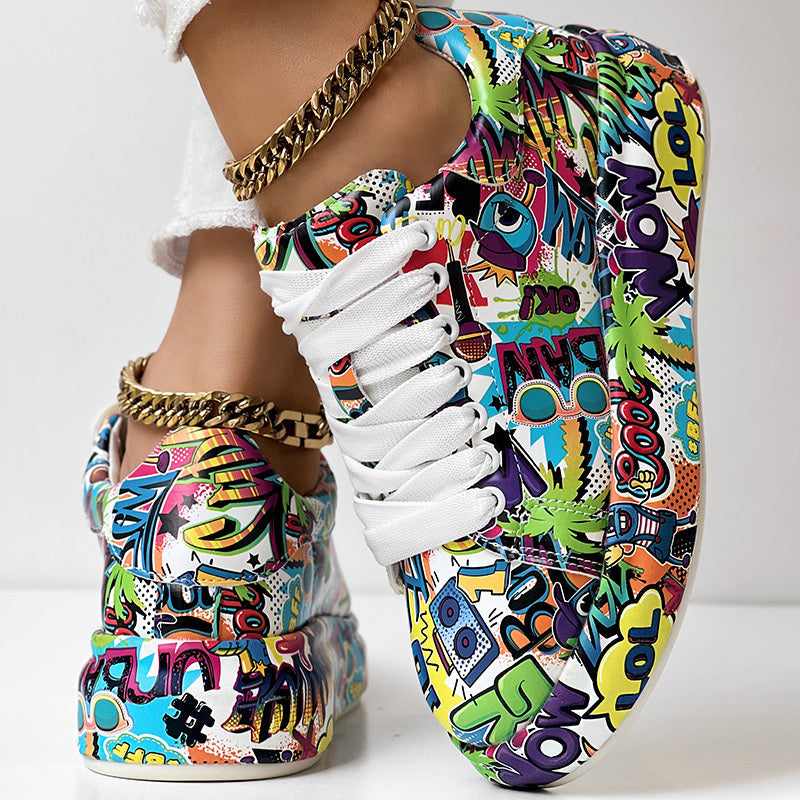 White lace-up sneakers featuring a vibrant, multicolored pop art graffiti print and gold chain ankle accessories.