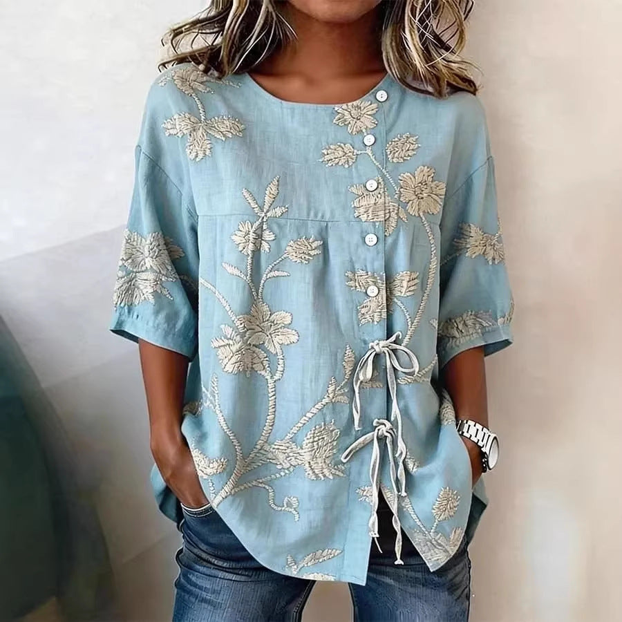 Light blue, short-sleeve linen blouse with cream floral embroidery, side button closure, and delicate tie details.