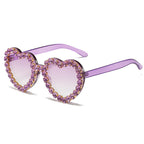Purple heart-shaped sunglasses with light purple gradient lenses and rhinestone-encrusted frames.