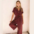 A woman wears a textured, short-sleeve maroon jumpsuit with a button-up V-neck and a matching tie-waist belt.