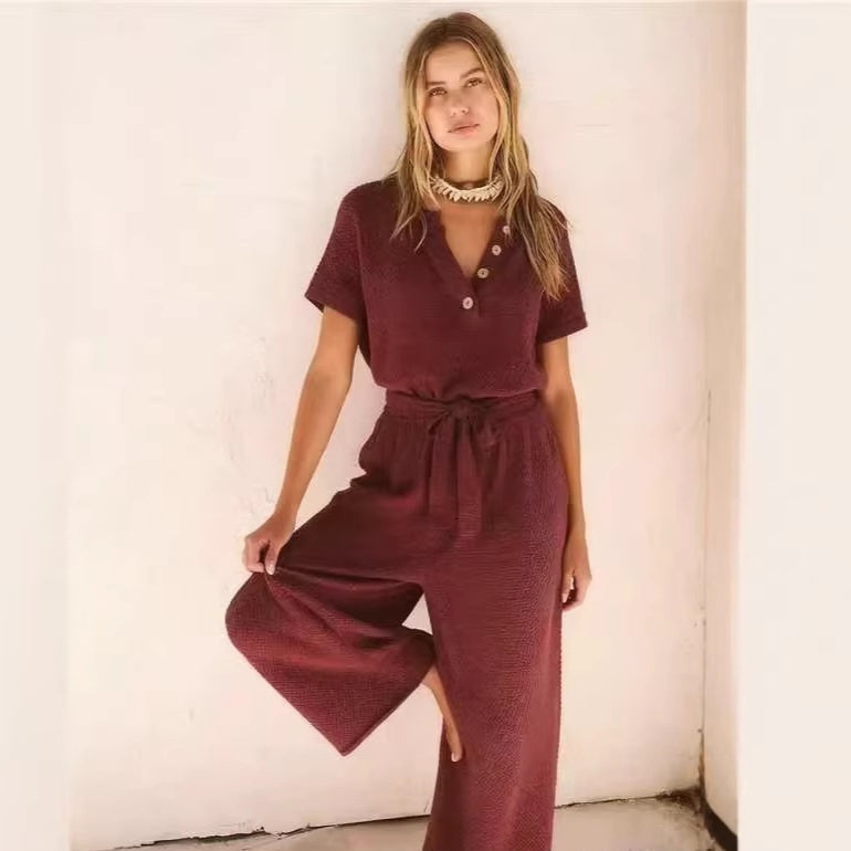 A woman wears a textured, short-sleeve maroon jumpsuit with a button-up V-neck and a matching tie-waist belt.