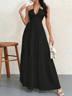 A woman wears a black, sleeveless halter maxi dress with a tiered skirt, holding a small white handbag.