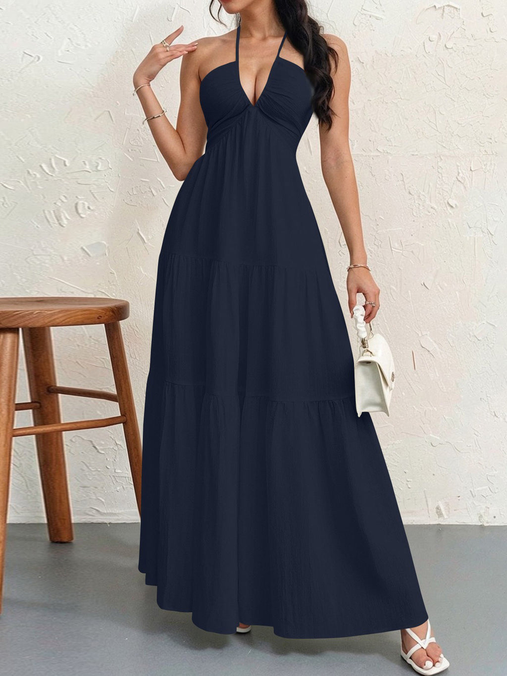 A woman wears a navy blue halter maxi dress with a deep V-neck and a tiered skirt, holding a small white handbag.
