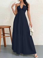 A woman wears a navy blue halter maxi dress with a deep V-neck and a tiered skirt, holding a small white handbag.