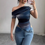 A woman wears a one-shoulder blue and black mesh top with a ruched, asymmetrical hem, paired with blue jeans.