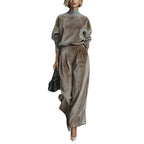 A woman wears a taupe velour high-neck long-sleeve top and matching wide-leg pleated trousers with a black handbag.