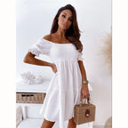 A woman wears a white, off-the-shoulder, smocked mini dress with puff sleeves and carries a woven rattan box bag.