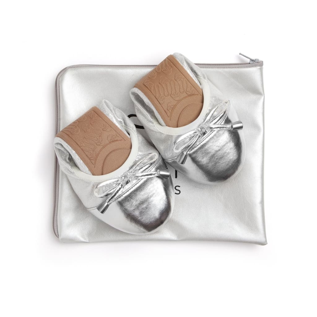 Pair of metallic silver ballet flats with front bows and tan soles, resting on a matching silver travel pouch.